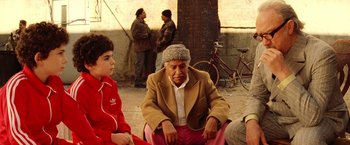 Movie still from “The Royal Tenenbaums” (2001), directed by Wes Anderson – An older man sitting next to a young boy in front of other people; Medium shot, High angle