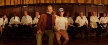 Movie still from “The Royal Tenenbaums” (2001), directed by Wes Anderson – A group of people sitting in a row of chairs; Medium shot, Low angle