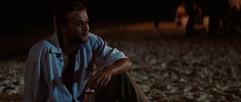 Movie still from “The Ruins” (2008), directed by Carter Smith – A man sitting on the ground holding a can of beer; Medium shot, Over the shoulder angle