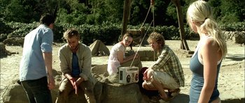Movie still from “The Ruins” (2008), directed by Carter Smith – A group of people sitting on a rock with a radio; Medium shot, Low angle