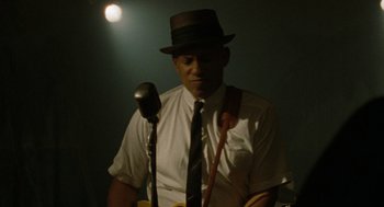 Movie still from “The Rum Diary” (2011), directed by Bruce Robinson – A man wearing a hat and a white shirt is playing a guitar; Medium shot, Low angle
