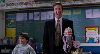 Movie still from “The Santa Clause” (1994), directed by John Pasquin – A man in a suit and tie holding a doll in a classroom; Medium shot, Over the shoulder angle
