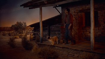 Movie still from “The Searchers” (1956), directed by John Ford – A man standing on a porch with a dog; Wide shot, Low angle