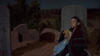 Movie still from “The Searchers” (1956), directed by John Ford – A young girl holding a banana sitting on the ground at night; Medium shot, High angle