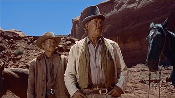 Movie still from “The Searchers” (1956), directed by John Ford – Two men in cowboy hats standing on a rocky hill; Medium shot, Low angle