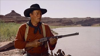 Movie still from “The Searchers” (1956), directed by John Ford – A man holding a gun while standing next to a body of water; Medium shot, Low angle
