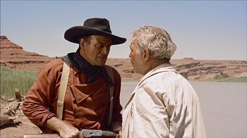 Movie still from “The Searchers” (1956), directed by John Ford – Two men talking to each other while holding a gun; Medium shot, Over the shoulder angle