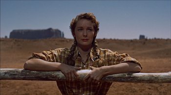 Movie still from “The Searchers” (1956), directed by John Ford – A woman in a plaid shirt leaning on a wooden fence; Medium shot, Low angle