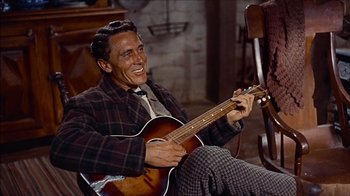 Movie still from “The Searchers” (1956), directed by John Ford – A man sitting in a chair holding a guitar; Medium shot, High angle