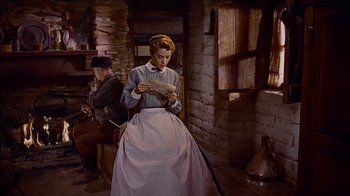 Movie still from “The Searchers” (1956), directed by John Ford – A woman sitting in front of a brick wall while a man stands next to her; Medium shot, Low angle