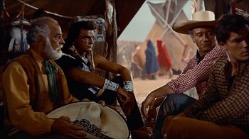Movie still from “The Searchers” (1956), directed by John Ford – A group of men sitting next to each other on the ground; Medium shot, Over the shoulder angle