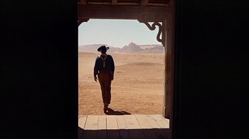 Movie still from “The Searchers” (1956), directed by John Ford – A man in a cowboy hat standing in front of an open door; Wide shot, Low angle