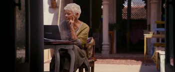 Movie still from “The Second Best Exotic Marigold Hotel” (2015), directed by John Madden – An older woman sitting at a table with a laptop computer in front of her; Medium shot, Over the shoulder angle