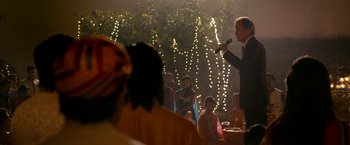 Movie still from “The Second Best Exotic Marigold Hotel” (2015), directed by John Madden – A man holding a microphone in front of a group of people; Medium shot, Over the shoulder angle