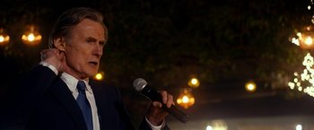 Movie still from “The Second Best Exotic Marigold Hotel” (2015), directed by John Madden – A man holding a microphone while standing in front of trees; Close Up shot, Over the shoulder angle