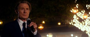 Movie still from “The Second Best Exotic Marigold Hotel” (2015), directed by John Madden – A man holding a microphone while standing in front of trees; Close Up shot, Over the shoulder angle