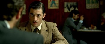 Movie still from “The Secret in Their Eyes” (2009), directed by Juan José Campanella – A man wearing a suit and tie sitting at a table in front of two other men; Medium shot, Over the shoulder angle