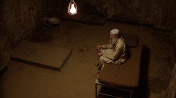 Movie still from “The Siege” (1998), directed by Edward Zwick – An older man sitting on a bed in a cave; Wide shot, High angle