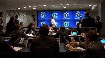 Movie still from “The Siege” (1998), directed by Edward Zwick – A group of people sitting in front of a large screen in a room; Wide shot, High angle