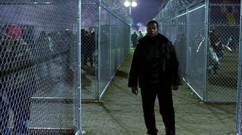 Movie still from “The Siege” (1998), directed by Edward Zwick – A man standing in front of a chain link fence at night; Wide shot, Over the shoulder angle