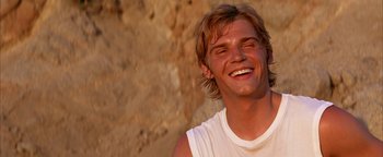 Movie still from “The Sisterhood of the Traveling Pants” (2005), directed by Ken Kwapis – A young man smiles brightly as he stands in front of a stone wall; Close Up shot, Over the shoulder angle