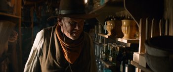 Movie still from “The Sisters Brothers” (2018), directed by Jacques Audiard – A man wearing a hat and a neck scarf looking at a shelf; Medium shot, Low angle