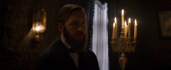 Movie still from “The Sisters Brothers” (2018), directed by Jacques Audiard – A man with a long beard standing in front of candles; Close Up shot, Over the shoulder angle