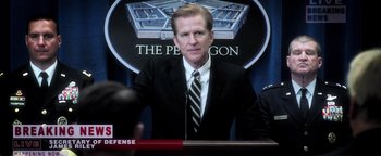 Movie still from “Sicario: Day of the Soldado” (2018), directed by Stefano Sollima – A man wearing a suit and tie sitting in front of a sign; Close Up shot, Over the shoulder angle