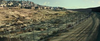 Movie still from “Sicario: Day of the Soldado” (2018), directed by Stefano Sollima – A wire fence in the middle of a field with a city in the background; Extreme Wide shot, High angle