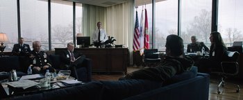 Movie still from “Sicario: Day of the Soldado” (2018), directed by Stefano Sollima – A man is sitting at a desk in front of two other men; Wide shot, Over the shoulder angle