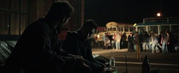 Movie still from “Sicario: Day of the Soldado” (2018), directed by Stefano Sollima – Two young men sitting on a bench at night with a bus in the background; Medium shot, Over the shoulder angle