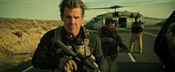 Movie still from “Sicario: Day of the Soldado” (2018), directed by Stefano Sollima – A man holding a gun while standing next to a helicopter; Medium shot, Over the shoulder angle