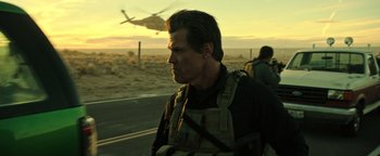 Movie still from “Sicario: Day of the Soldado” (2018), directed by Stefano Sollima – A man standing on the side of a road next to a helicopter; Medium shot, Over the shoulder angle
