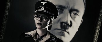 A woman wearing a hat and glasses in front of a picture of adolf hitler