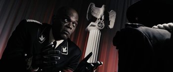 Movie still from “The Spirit” (2008), directed by Frank Miller – A man in a black suit and white shirt is standing in front of a statue of an eagle; Close Up shot, Over the shoulder angle