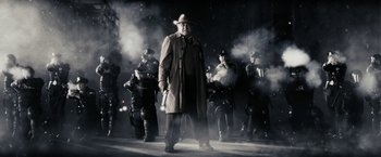 Movie still from “The Spirit” (2008), directed by Frank Miller – A man wearing a hat and a trench coat standing in front of a group of people; Medium shot, Low angle
