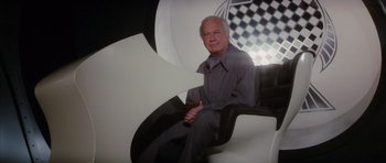 Movie still from “The Spy Who Loved Me” (1977), directed by Lewis Gilbert – An older man sitting on a chair in front of a checkerboard wall; Medium shot, Low angle