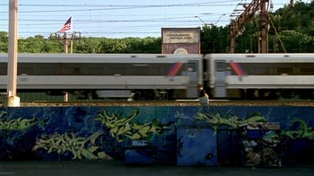 Movie still from “The Station Agent” (2003), directed by Tom McCarthy – A train traveling down train tracks next to a street; Extreme Wide shot, High angle