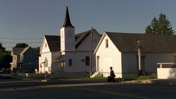 Movie still from “The Station Agent” (2003), directed by Tom McCarthy – A person sitting on the side of a road near a church; Extreme Wide shot, Low angle