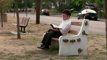 Movie still from “The Station Agent” (2003), directed by Tom McCarthy – A man sitting on top of a wooden bench reading a book; Wide shot, Low angle