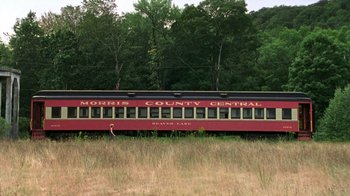 Movie still from “The Station Agent” (2003), directed by Tom McCarthy – A red and black passenger train traveling through a field; Extreme Wide shot, Low angle
