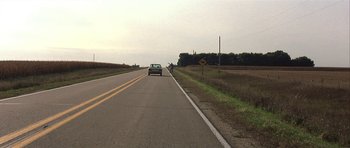 Movie still from “The Straight Story” (1999), directed by David Lynch – A car driving down a road near a grassy field; Extreme Wide shot, High angle