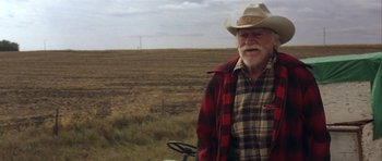 Movie still from “The Straight Story” (1999), directed by David Lynch – An older man wearing a cowboy hat standing next to a tractor; Medium shot, Over the shoulder angle