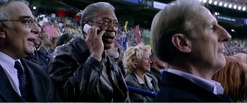Movie still from “The Sum of All Fears” (2002), directed by Phil Alden Robinson – An older man talking on a cell phone in front of an audience; Medium shot, Low angle