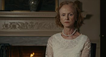 Movie still from “The Taking of Deborah Logan” (2014), directed by Adam Robitel – An older woman wearing a white dress and a pearl necklace in front of a fireplace; Close Up shot, Over the shoulder angle