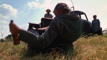 Movie still from “The Texas Chain Saw Massacre” (1974), directed by Tobe Hooper – A man sitting in the grass using a computer; Wide shot, Low angle