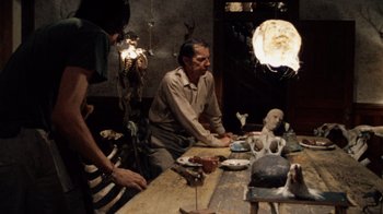 Movie still from “The Texas Chain Saw Massacre” (1974), directed by Tobe Hooper – A man sitting at a dining table with other people around him; Wide shot, Over the shoulder angle