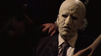 Movie still from “The Texas Chain Saw Massacre” (1974), directed by Tobe Hooper – A man in a suit and tie with a mask on his face; Close Up shot, Over the shoulder angle