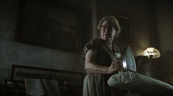 Movie still from “The Texas Chainsaw Massacre” (2003), directed by Marcus Nispel – An older woman holding a iron in a dark room; Medium shot, Low angle