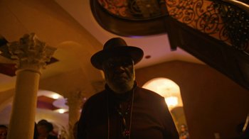 Movie still from “Full Circle” (2023), directed by Steven Soderbergh – An older man wearing a hat standing in front of a staircase; Medium shot, Low angle